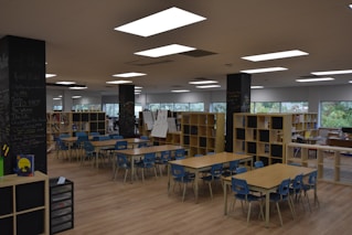 A spacious classroom or learning area filled with several wooden tables and blue chairs arranged neatly. Bookshelves line the room, filled with books and educational materials. The walls feature large windows that provide natural light. Chalkboards are visible on some pillars, displaying handwritten notes and drawings.