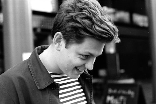A black and white photograph of a person with short hair, looking down, and smiling. They are wearing a striped shirt under a jacket. The background is blurred, suggesting an outdoor setting with indiscernible details.