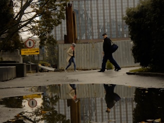 Two people are walking on a wet street, with their reflections visible in a large puddle. A tall building with a grid-like glass facade is in the background, partially obscured by a construction fence. Trees on the left and right frame the scene. A sign with Polish text and a pedestrian icon is visible.