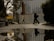 Two people are walking on a wet street, with their reflections visible in a large puddle. A tall building with a grid-like glass facade is in the background, partially obscured by a construction fence. Trees on the left and right frame the scene. A sign with Polish text and a pedestrian icon is visible.