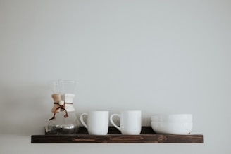 A clean, minimalist display of handcrafted ceramic mugs on a simple wooden shelf.