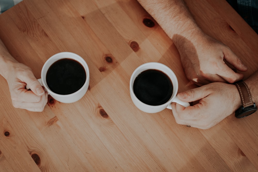 Zwei Personen halten weiße Tassen gefüllt mit Kaffee auf brauner Oberfläche