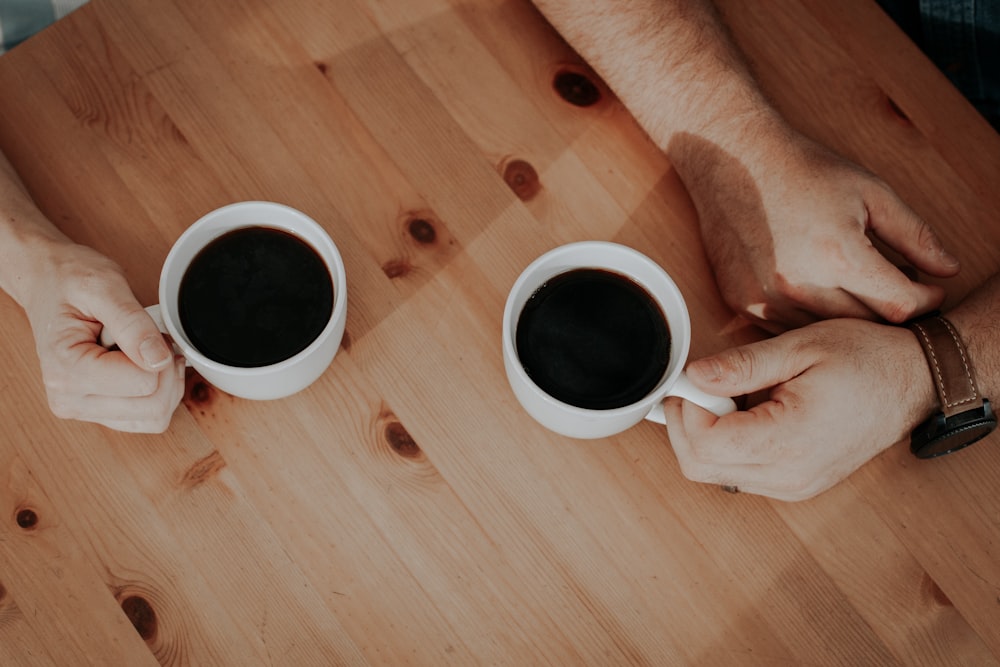 duas pessoas segurando canecas brancas cheias de café na superfície marrom