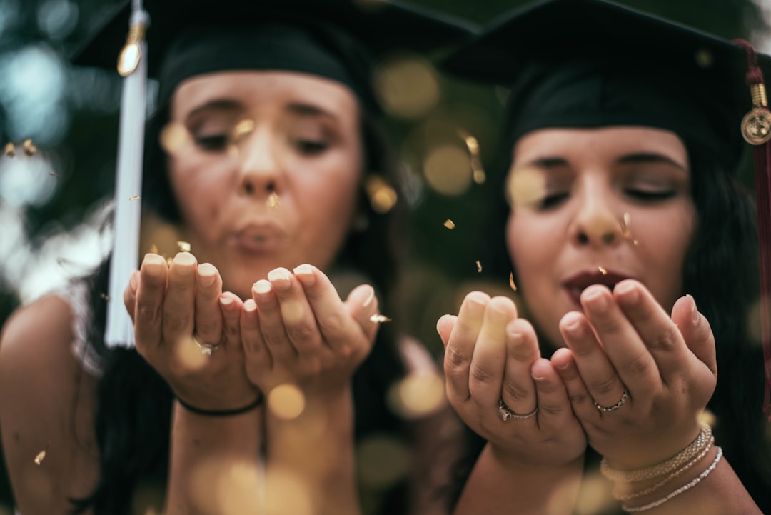 Two woman wearing graduation caps open their palms and blow confetti as an act of celebration