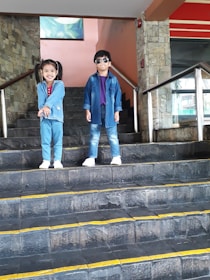 Two children stand on a set of stone steps. The child on the left is smiling, wearing a blue jacket over a red shirt and white sneakers. The child on the right stands confidently with hands by their sides, wearing sunglasses, a blue jacket, a purple shirt, and white sneakers.