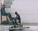 A shoe shiner is polishing the shoes of a man seated on a high wooden chair against a tiled wall. The shoe shiner is hunched over, focused on his work, while the seated man looks towards the camera wearing a red cap and a blue shirt. Several pairs of shoes are placed on a small wooden platform on the ground.