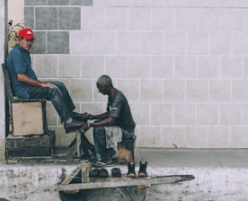 A shoe shiner is polishing the shoes of a man seated on a high wooden chair against a tiled wall. The shoe shiner is hunched over, focused on his work, while the seated man looks towards the camera wearing a red cap and a blue shirt. Several pairs of shoes are placed on a small wooden platform on the ground.
