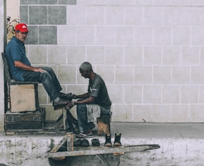 A shoe shiner is polishing the shoes of a man seated on a high wooden chair against a tiled wall. The shoe shiner is hunched over, focused on his work, while the seated man looks towards the camera wearing a red cap and a blue shirt. Several pairs of shoes are placed on a small wooden platform on the ground.