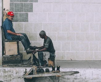 A shoe shiner is polishing the shoes of a man seated on a high wooden chair against a tiled wall. The shoe shiner is hunched over, focused on his work, while the seated man looks towards the camera wearing a red cap and a blue shirt. Several pairs of shoes are placed on a small wooden platform on the ground.