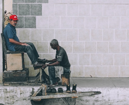 A shoe shiner is polishing the shoes of a man seated on a high wooden chair against a tiled wall. The shoe shiner is hunched over, focused on his work, while the seated man looks towards the camera wearing a red cap and a blue shirt. Several pairs of shoes are placed on a small wooden platform on the ground.