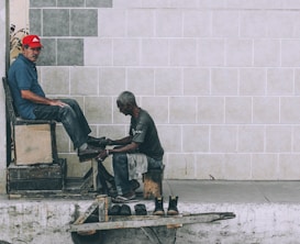A shoe shiner is polishing the shoes of a man seated on a high wooden chair against a tiled wall. The shoe shiner is hunched over, focused on his work, while the seated man looks towards the camera wearing a red cap and a blue shirt. Several pairs of shoes are placed on a small wooden platform on the ground.