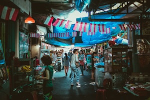 A bustling market scene with numerous people walking along a narrow street lined with stalls on either side. Various goods are displayed, and vibrant fabrics are strung above, providing shade. The atmosphere is lively and colorful, suggesting a local marketplace ambiance.