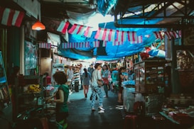 A bustling market scene with numerous people walking along a narrow street lined with stalls on either side. Various goods are displayed, and vibrant fabrics are strung above, providing shade. The atmosphere is lively and colorful, suggesting a local marketplace ambiance.