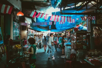 A bustling market scene with numerous people walking along a narrow street lined with stalls on either side. Various goods are displayed, and vibrant fabrics are strung above, providing shade. The atmosphere is lively and colorful, suggesting a local marketplace ambiance.