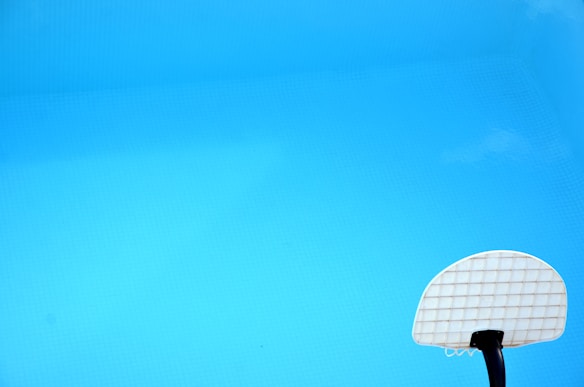A swimming pool with clear blue water featuring a white basketball hoop in the lower right corner.