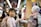 A young child with a backpack holds hands with a humanoid robot adorned with pink flowers and equipped with a tablet on its chest. They are standing in what appears to be a busy market or shopping area with bright, blurred lights and people in the background.