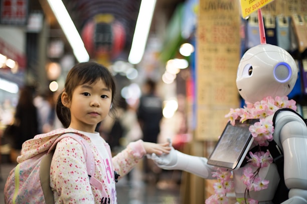 A young child with a backpack holds hands with a humanoid robot adorned with pink flowers and equipped with a tablet on its chest. They are standing in what appears to be a busy market or shopping area with bright, blurred lights and people in the background.