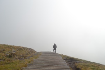 A solitary figure walks along a wooden pathway surrounded by mist and fog. The landscape is sparse, with grass and rocks lining the sides of the path, giving a sense of isolation and tranquility. The horizon is obscured by the fog, creating an ethereal atmosphere.