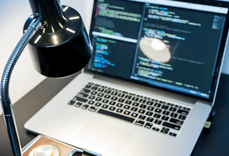 A workspace with a laptop displaying code, featuring a notable dark and minimal design. The screen shows multiple lines of colorful code against a dark background, indicating software development in progress. A sleek black desk lamp with a chrome adjustable arm illuminates part of the desk. The setting suggests a focused and tech-savvy environment for coding or programming tasks.