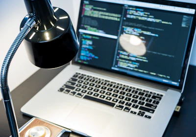 A workspace with a laptop displaying code, featuring a notable dark and minimal design. The screen shows multiple lines of colorful code against a dark background, indicating software development in progress. A sleek black desk lamp with a chrome adjustable arm illuminates part of the desk. The setting suggests a focused and tech-savvy environment for coding or programming tasks.