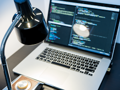 A workspace with a laptop displaying code, featuring a notable dark and minimal design. The screen shows multiple lines of colorful code against a dark background, indicating software development in progress. A sleek black desk lamp with a chrome adjustable arm illuminates part of the desk. The setting suggests a focused and tech-savvy environment for coding or programming tasks.