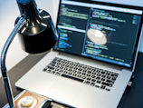 A workspace with a laptop displaying code, featuring a notable dark and minimal design. The screen shows multiple lines of colorful code against a dark background, indicating software development in progress. A sleek black desk lamp with a chrome adjustable arm illuminates part of the desk. The setting suggests a focused and tech-savvy environment for coding or programming tasks.