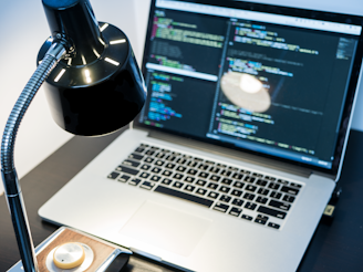 A workspace with a laptop displaying code, featuring a notable dark and minimal design. The screen shows multiple lines of colorful code against a dark background, indicating software development in progress. A sleek black desk lamp with a chrome adjustable arm illuminates part of the desk. The setting suggests a focused and tech-savvy environment for coding or programming tasks.