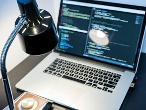 A workspace with a laptop displaying code, featuring a notable dark and minimal design. The screen shows multiple lines of colorful code against a dark background, indicating software development in progress. A sleek black desk lamp with a chrome adjustable arm illuminates part of the desk. The setting suggests a focused and tech-savvy environment for coding or programming tasks.