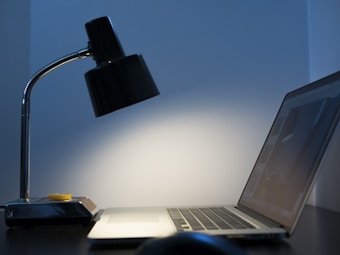 A dimly lit workspace featuring a sleek black desk lamp and an open laptop positioned on a dark surface. The lamp is turned on, casting a soft glow on the wall behind. The ambiance is minimalistic and modern.