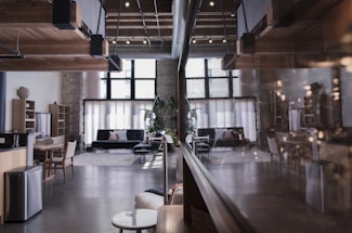 A spacious, modern interior with a loft design. The room features wooden ceiling beams, large windows that let in ample natural light, and contemporary furnishings including sofas and a dining table. The atmosphere is enhanced by a mix of exposed brick and sleek elements, such as a polished floor and minimalistic decor.