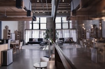 A spacious, modern interior with a loft design. The room features wooden ceiling beams, large windows that let in ample natural light, and contemporary furnishings including sofas and a dining table. The atmosphere is enhanced by a mix of exposed brick and sleek elements, such as a polished floor and minimalistic decor.