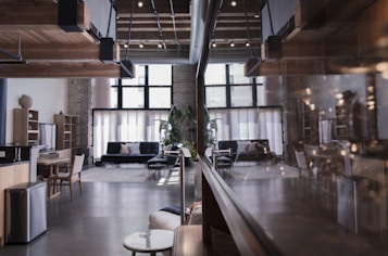 A spacious, modern interior with a loft design. The room features wooden ceiling beams, large windows that let in ample natural light, and contemporary furnishings including sofas and a dining table. The atmosphere is enhanced by a mix of exposed brick and sleek elements, such as a polished floor and minimalistic decor.