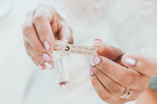Hands with pink geometric nail art hold a small glass vial sealed with a cork. A paper tag attached to the vial reads 'We tied the knot, now take a shot'.