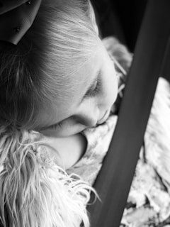 A young child with a bow in their hair is asleep, leaning against a fluffy blanket while secured by a seatbelt. The image is captured in black and white, highlighting soft textures and peaceful expression.