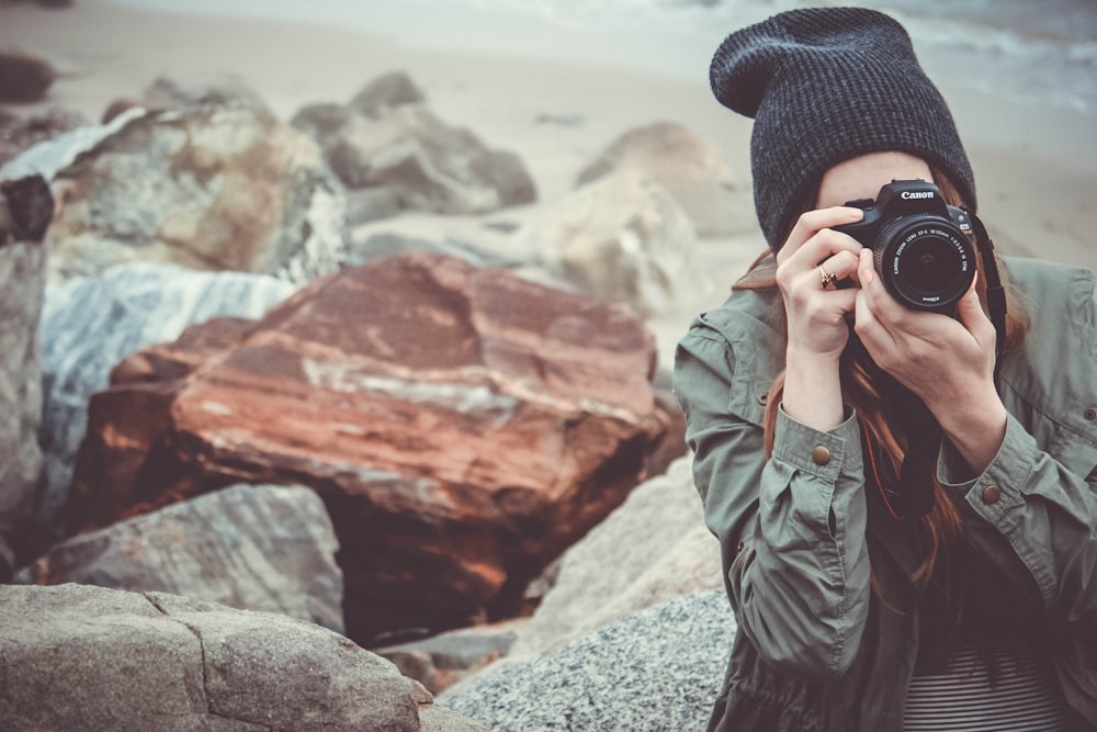 회색 재킷과 검은 니트 모자를 쓴 여자가 낮에 큰 바위 근처에서 검은 캐논 DSLR 카메라를 들고 있습니다.