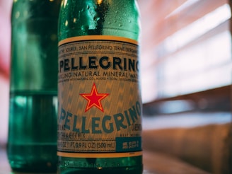 A green glass bottle with a prominent label featuring the name Pellegrino and a red star. The bottle contains sparkling natural mineral water and the background shows blurred horizontal lines, likely representing window blinds with sunlight filtering through.