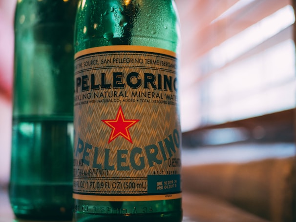 A green glass bottle with a prominent label featuring the name Pellegrino and a red star. The bottle contains sparkling natural mineral water and the background shows blurred horizontal lines, likely representing window blinds with sunlight filtering through.