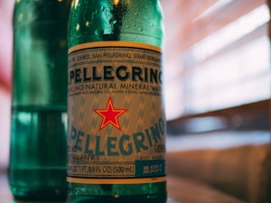 A green glass bottle with a prominent label featuring the name Pellegrino and a red star. The bottle contains sparkling natural mineral water and the background shows blurred horizontal lines, likely representing window blinds with sunlight filtering through.