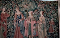 A richly detailed tapestry features four figures in medieval attire, surrounded by lush foliage, birds, and flowers. The individuals are dressed in ornate garments with patterns and textures that suggest wealth and nobility. The background is densely populated with intricate depictions of leaves and blossoms.