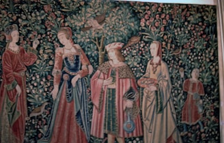 A richly detailed tapestry features four figures in medieval attire, surrounded by lush foliage, birds, and flowers. The individuals are dressed in ornate garments with patterns and textures that suggest wealth and nobility. The background is densely populated with intricate depictions of leaves and blossoms.