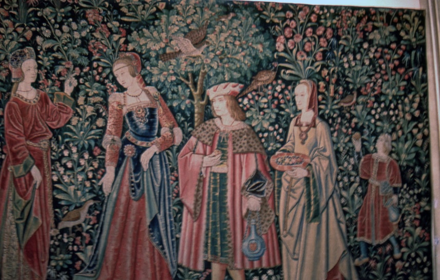 A richly detailed tapestry features four figures in medieval attire, surrounded by lush foliage, birds, and flowers. The individuals are dressed in ornate garments with patterns and textures that suggest wealth and nobility. The background is densely populated with intricate depictions of leaves and blossoms.