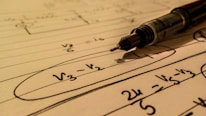 A mechanical pencil rests on a notebook filled with handwritten mathematical equations. The formulas include terms such as V3, V2 with elements circled and lines drawn across the page. The use of pen and paper suggests an academic or study setting.