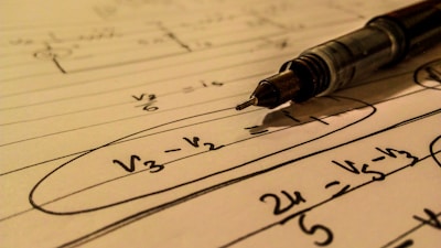 A mechanical pencil rests on a notebook filled with handwritten mathematical equations. The formulas include terms such as V3, V2 with elements circled and lines drawn across the page. The use of pen and paper suggests an academic or study setting.