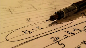 A mechanical pencil rests on a notebook filled with handwritten mathematical equations. The formulas include terms such as V3, V2 with elements circled and lines drawn across the page. The use of pen and paper suggests an academic or study setting.