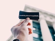 A hand holding up a black card with white text that reads 'creativity doesn't need limits.' The background features an out-of-focus building with tall columns.