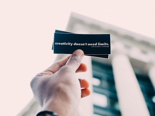 A hand holding up a black card with white text that reads 'creativity doesn't need limits.' The background features an out-of-focus building with tall columns.