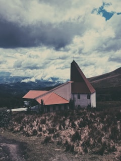 A quaint church with a dark brown steeple and red-tiled roof is nestled in a rustic landscape with tall, golden grasses. The backdrop features rolling mountains under a dramatic, cloud-filled sky.
