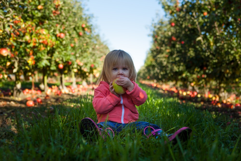 girl sitting on green grass field holding green fruit during daytime