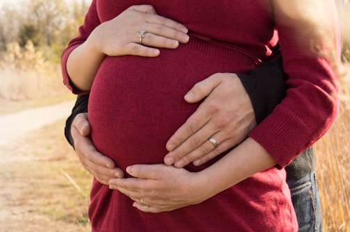 Two pairs of hands gently rest on a pregnant belly, symbolizing care and expectation. The person is wearing a red sweater, and there are wedding rings visible on some of the fingers. The background is blurred, with hints of autumnal colors.
