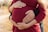 Two pairs of hands gently rest on a pregnant belly, symbolizing care and expectation. The person is wearing a red sweater, and there are wedding rings visible on some of the fingers. The background is blurred, with hints of autumnal colors.